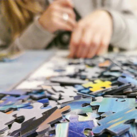 A close up of puzzle pieces with someone putting pieces in place across the table.