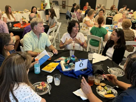 A room full of adults sitting at round tables listening to different authors while eating a light lunch
