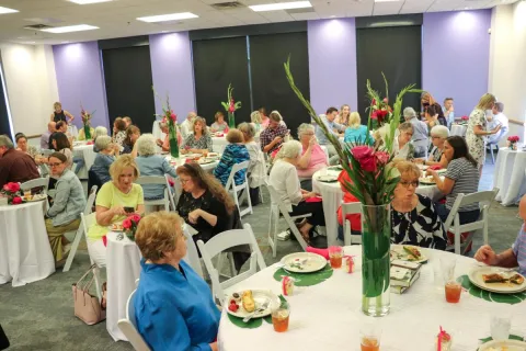 A lunch reception with flower centerpieces and groups sitting at tables enjoying food with copies of books on tables